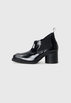 Bottine en cuir verni noir avec un talon épais, des panneaux élastiques sur les côtés, un bout arrondi et une languette à l'arrière pour un enfilage facile.