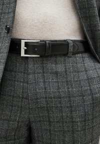 Pantalon à carreaux gris avec un motif subtil, associé à une ceinture en cuir noir lisse avec une boucle en métal argenté. Doux pull beige en dessous.