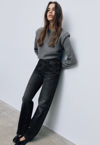 Szary sweter z dzianiny z wypełnionymi ramionami, połączony z czarnymi, wysokim stanem, prostymi spodniami dżinsowymi. Modelka opiera się o jasną ścianę, mając na sobie czarne buty.