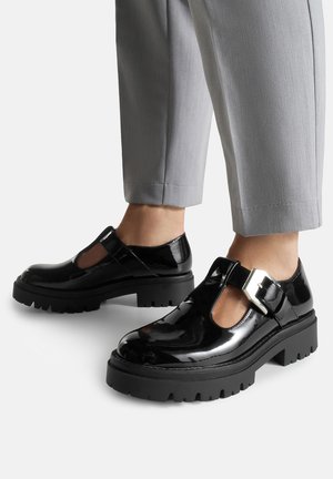 Scarpe Mary Jane in vernice nera con suole carrarmato chunky e dettaglio fibbia dorata, indossate con pantaloni grigi chiari a vita alta.