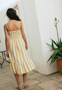 Robe sans bretelles, jaune et blanche à rayures, avec un buste ajusté et un ourlet à volants évasé. Le mannequin se tient contre un mur, des sandales visibles.