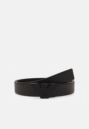 Ceinture en cuir noir avec une finition texturée et une grande boucle en forme d’aigle, posée enroulée sur un fond blanc.