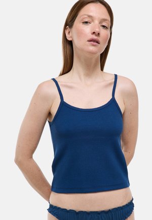 Femme aux cheveux raides portant un caraco bleu marine ajusté et une lingerie assortie à volants sur un fond clair uni.