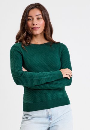 Pull en tricot vert avec une texture en nid d'abeille, col rond et manches longues. Poignets et ourlet côtelés ; associé à un jean bleu clair.