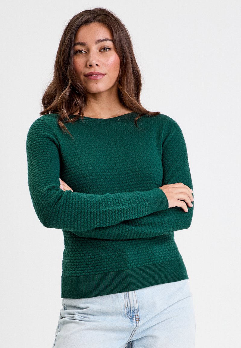 Pull en tricot vert avec une texture en nid d'abeille, col rond et manches longues. Poignets et ourlet côtelés ; associé à un jean bleu clair.