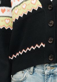 Μαύρο πλεκτό ζακέτα με πορτοκαλί και λευκά σχέδια καρδιών, με ζιγκ ζαγκ περίγραμμα, μαύρα κουμπιά με τη φίρμα "Moschino Jeans", συνδυασμένο με ανοιχτόχρωμα γαλάζια τζιν.