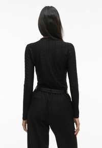 Musta pitkähihainen paita, jossa on tekstuuri, yhdistetty korkeavyötäröisiin mustiin housuihin. Asusteessa on kapea vyö ja huomaamattomat taskuyksityiskohdat.