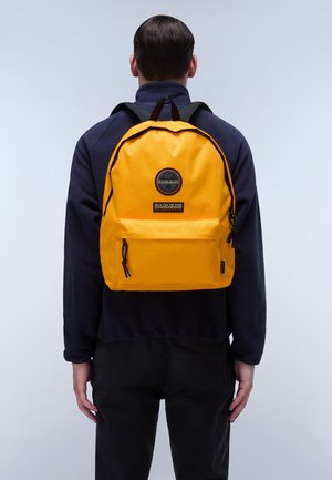 Zaino giallo dalla forma arrotondata, con una tasca frontale e accessori in metallo neri. Realizzato in tessuto resistente, indossato da una persona.