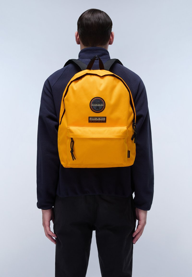 Sac à dos jaune avec une forme arrondie, doté d'une poche avant et d'accents en métal noir. Fabriqué en tissu résistant, porté par une personne.