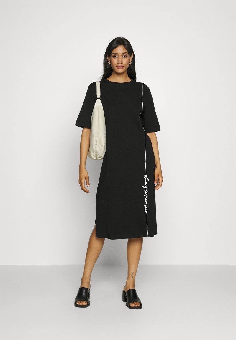 Mujer vestida con un vestido midi negro con texto blanco en vertical, sandalias de tacón negras, de pie con un bolso beige al hombro sobre un fondo sencillo.