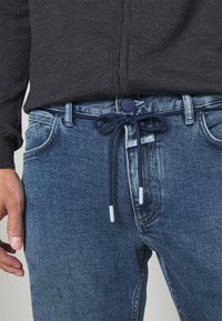 Mörkgrå zip-up tröja ovanpå blå denimjeans med dragsko i midjan. Jeansen har en klassisk femfickdesign och en lätt blekning.