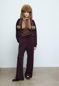 Brauner, mit Fell gefütterter Hut, weinroter Reißverschluss-Hoodie mit Pompons, braunes T-Shirt, weite weinrote Hose und dunkle Hausschuhe. Neutraler Hintergrund.