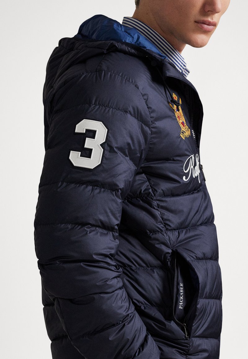 Man wearing navy blue padded jacket with hood, embroidered number "3" on sleeve, crest on chest, and partially unzipped pocket labeled "PACKABLE".