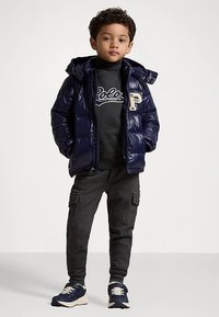 Νεαρός αγόρι στέκεται με τα χέρια στις τσέπες φορώντας ένα ναυτικό σακάκι, σκούρο γκρι φούτερ Polo, cargo παντελόνι και ναυτικά αθλητικά παπούτσια.