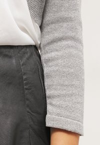 Bras dans une manche en maille grise à côté d'une jupe en cuir noir et d'un chemisier blanc rentré, mettant en valeur les textures des tissus et la coupe des vêtements.