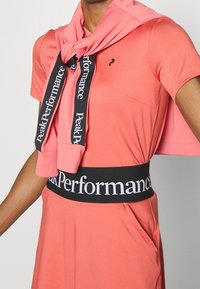 Abito atletico rosa con maniche corte, caratterizzato da una tasca anteriore, abbinato a una fascia elastica nera e bianca con marchio e un foulard coordinato.