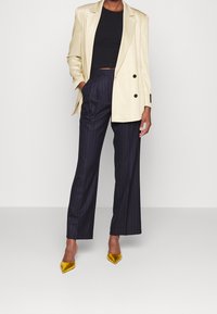 Blazer beige de doble botonadura sobre un top negro corto, combinado con pantalones de pierna ancha de rayas oscuras y tacones amarillos de satén en punta.