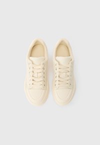 Kreemjad madala varrega tossud, millel on paelad, ümarad ninad ja isolaatoriga kraed, asetatud valge taustaga kõrvuti.