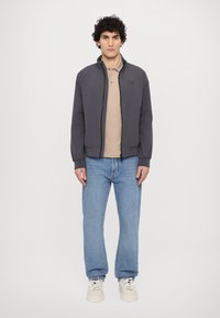 Grå zip-up jakke med høy krage, tan polo skjorte under, lyseblå rett-leg jeans, og hvite joggesko. Jevn stofftekstur.