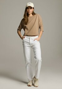 Beżowa bluzka o luźnym kroju z trzema ćwiartkami rękawów, białe dżinsy straight-leg i beżowe trampki. Kapelusz w kremowym kolorze z logo.