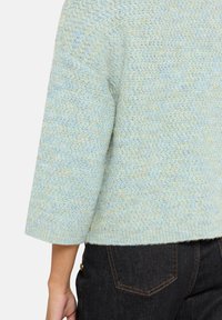 Jasnoniebieski sweter dziany z teksturowanym wzorem, rękawami do połowy przedramienia i krótkim dołem, zestawiony z ciemnymi jeansami.