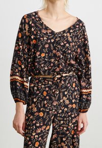 Haut à manches longues, décolleté en V, de couleur noire avec un motif floral dans des teintes d'orange et de rose. Caractérisé par des poignets élastiques et un détail à nouer à la taille.