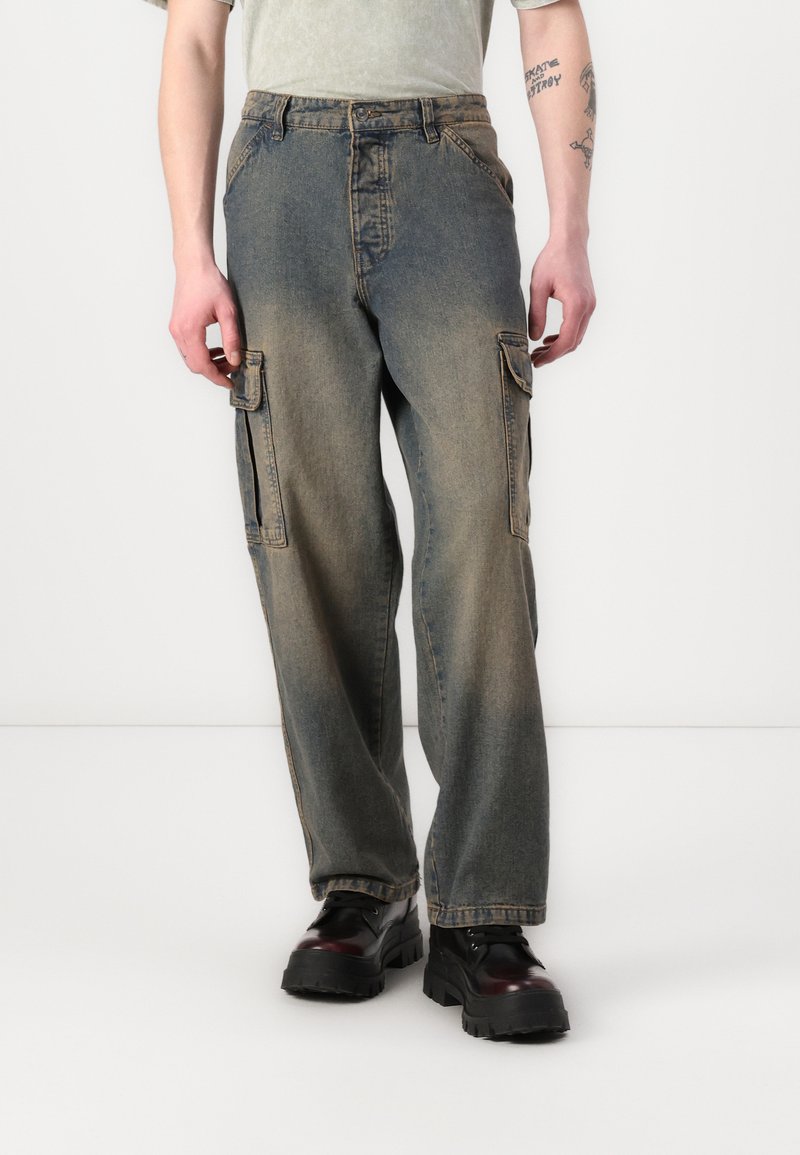 Denim-Cargo-Hosen in verblasstem Blau mit lockerer Passform, ausgestattet mit seitlichen Taschen und sichtbaren Nähten. Kombiniert mit klobigen schwarzen Stiefeln.