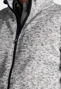 Close-up van een persoon in een grijs en zwart gespetterde ritstrui over een donker overhemd met een gele knoop.