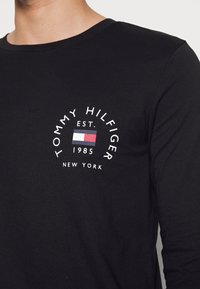 Musta pitkähihainen paita pehmeästä materiaalista, jossa on pyöreä logo, jossa lukee "TOMMY HILFIGER", lippumallinen muotoilu ja "EST. 1985 NEW YORK" valkoisella tekstillä.