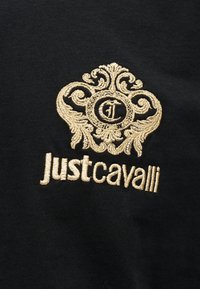 Χρυσό διακοσμητικό έμβλημα με τα αρχικά "JC" και το κείμενο "Just Cavalli" κεντημένα σε μαύρο ύφασμα.