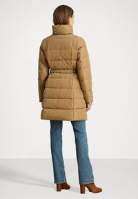 Kávészínű puffertakaró, magas gallérral, rugalmas derékkal és öv részlettel, valamint steppelt mintával. Kék farmerral és barna sarkú csizmával párosítva.