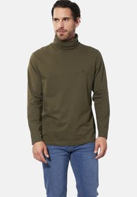 Grünes langärmliges Rollkragenshirt aus weichem Material. Hat eine schmale Passform und ein kleines Logo auf der Brust. Kombiniert mit blauen Jeans.