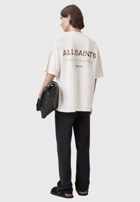 Camiseta de gran tamaño en color crema con el gráfico "ALLSAINTS" en letras coloridas en la parte posterior. Bolsa negra en la mano; combinada con pantalones negros y zapatos sin cordones.