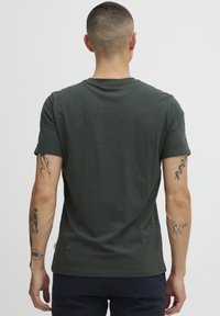 Camiseta de algodón de color verde oscuro con mangas cortas, cuello redondo y una textura suave. Tatuajes notables visibles en los brazos.