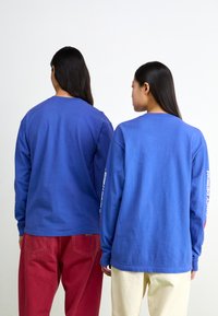 Langärmlige blaue Baumwoll-T-Shirts, die von zwei Personen getragen werden, mit bedrucktem Text auf dem linken Ärmel. Rote und beige Hosen sind darunter sichtbar.