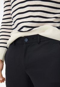 Zwarte broek met een gladde textuur, voorzien van een knoopsluiting, riemlussen en zijzakken, gecombineerd met een gestreepte gebreide trui.