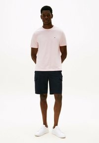 Jasnoróżowa bawełniana koszulka z krótkim rękawem i małym logo, zestawiona z granatowymi szortami cargo i białymi trampkami.