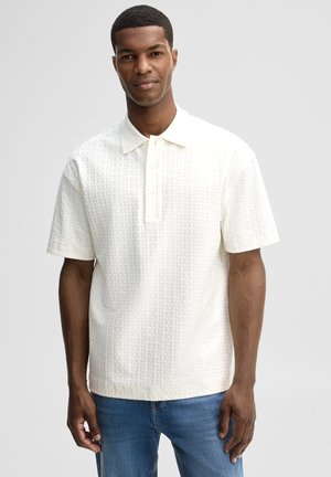 Uomo che indossa una polo bianca a maniche corte con tessuto strutturato e jeans blu, in piedi davanti a uno sfondo grigio chiaro uniforme.
