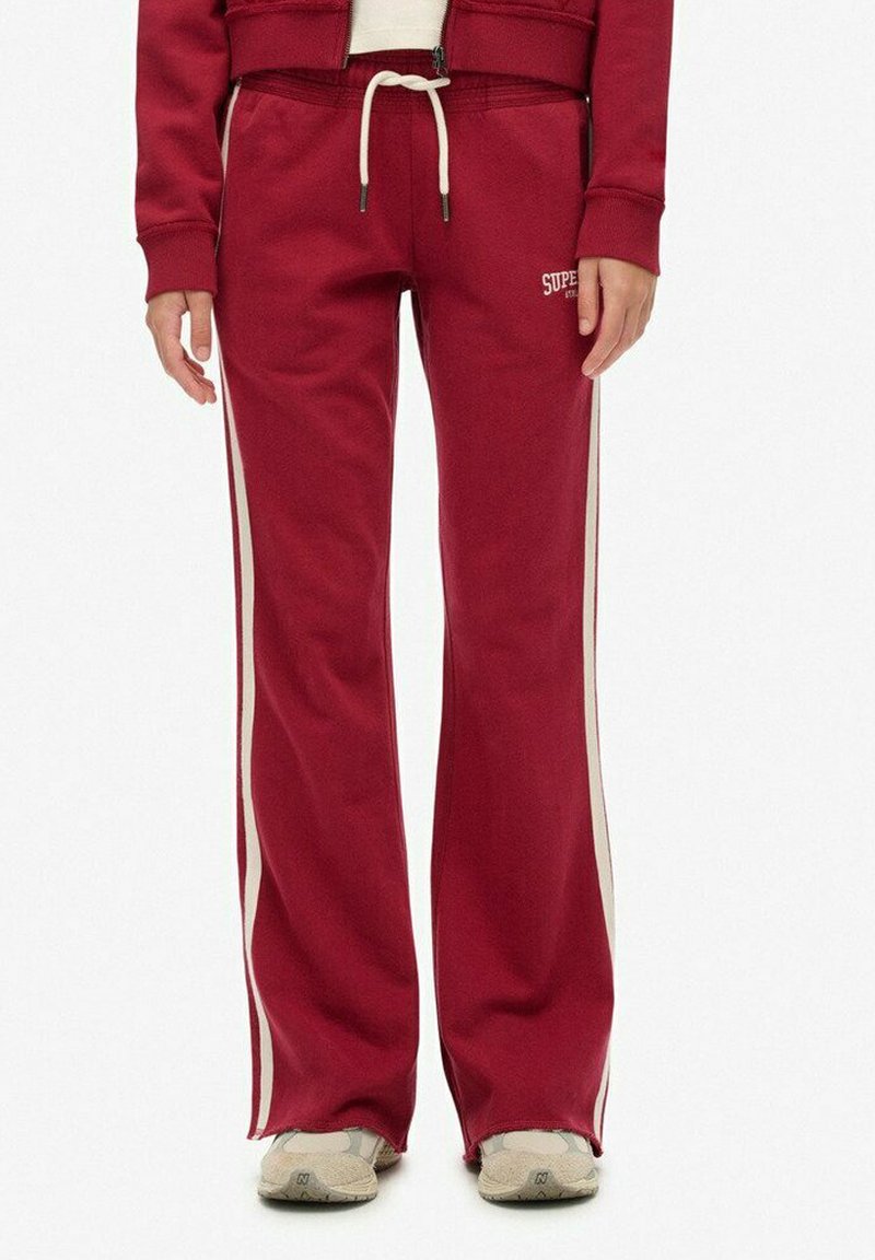 Red sweatpants made of soft fabric with a drawstring waist, featuring cream side stripes and a logo on the left thigh. Flared design.