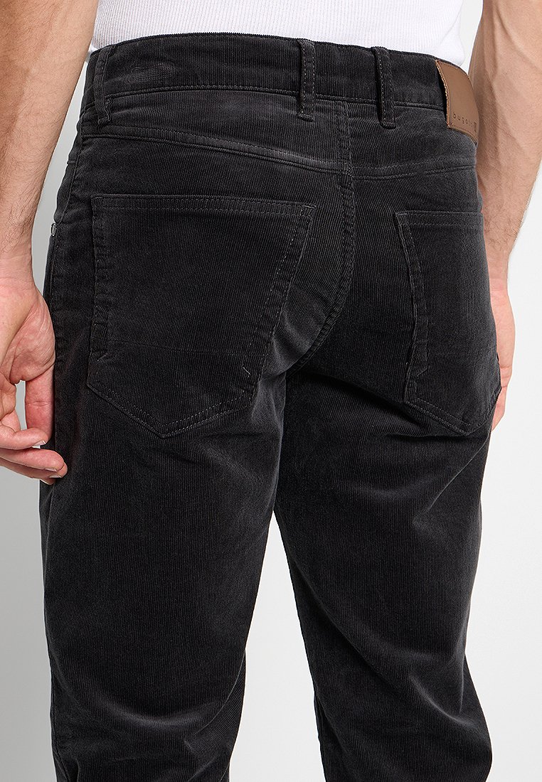 Mann in schwarzen Cordhosen und weißem Hemd, gezeigt von der Taille bis zur Mitte des Oberschenkels, mit sichtbaren Gesäßtaschen und Gürtelschlaufen.