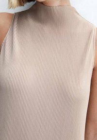 Top sin mangas de color beige claro con un diseño de cuello alto, que presenta una textura acanalada que recorre verticalmente la tela.