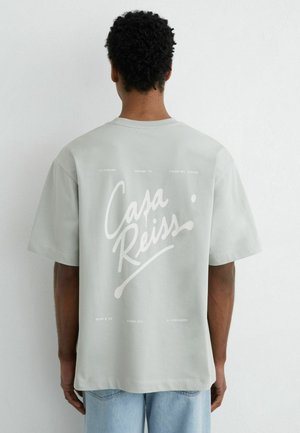 Homme aux cheveux courts et bouclés portant un t-shirt oversize gris clair avec le texte blanc « Casa Reiss » dans le dos, associé à un jean bleu clair.