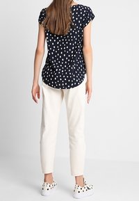 Granatowa bluzka w kropki z krótkimi rękawami, zestawiona z kremowymi, zwężanymi spodniami oraz białymi wsuwanymi sneakersami w czarne kropki. Gładka faktura materiału.