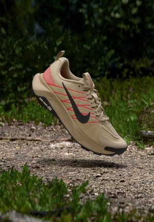 Μπεζ παπούτσι τρεξίματος για μονοπάτια της Nike με μαύρο σήμα και κόκκινες λεπτομέρειες που αιωρείται πάνω από ένα χαλικόδρομο σε πράσινο εξωτερικό περιβάλλον.