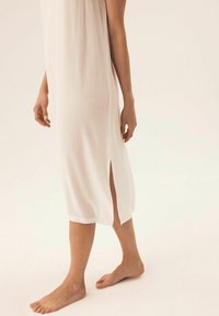 Długa, biała sukienka z teksturowanej tkaniny, z bocznym rozcięciem i luźnym krojem. Sukienka sięga poniżej kolana, odsłaniając nagie stopy.