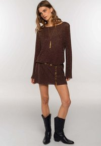 Top de punto marrón brillante de manga larga combinado con una mini falda a juego, que presenta un acento de cinturón circular dorado. Las botas cowboy negras completan el look.