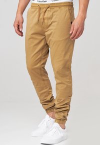 Hellbraune Jogginghosen mit elastischem Bund und Kordelzug. Ausgestattet mit Bündchen an den Knöcheln und Seitentaschen, kombiniert mit weißen Turnschuhen.