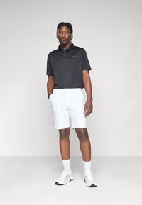 Svart polo med krage og liten logo, kombinert med hvite shorts og hvite joggesko. Antrekket er uformelt og atletisk i stil.
