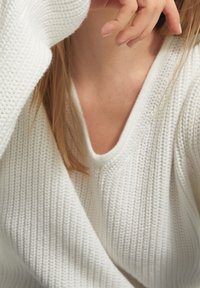 Biały sweter dziany z dekoltem w szpic, z teksturowanym wzorem. Miękki materiał, luźne rękawy oraz minimalne detale.
