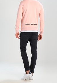 Vaaleanpunainen huppari, jossa on "CRIMINAL DAMAGE" -teksti alareunassa, yhdistettynä mustiin kapeisiin farkkuihin ja valkoisiin lenkkareihin.